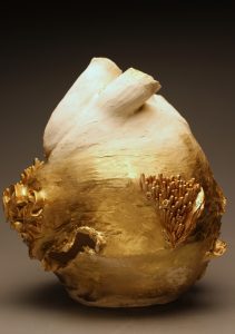 A sculpture of an anatomical heart which is white at the top but fades into gold at the bottom. At the midpoint, organic forms rise from the surface that resemble coral.