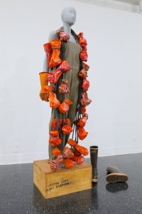 Life-sized figure standing on a wooden box wearing chest-high hip boots and rubber gloves. It is draped in a garland of dozens of red rubber gloves.